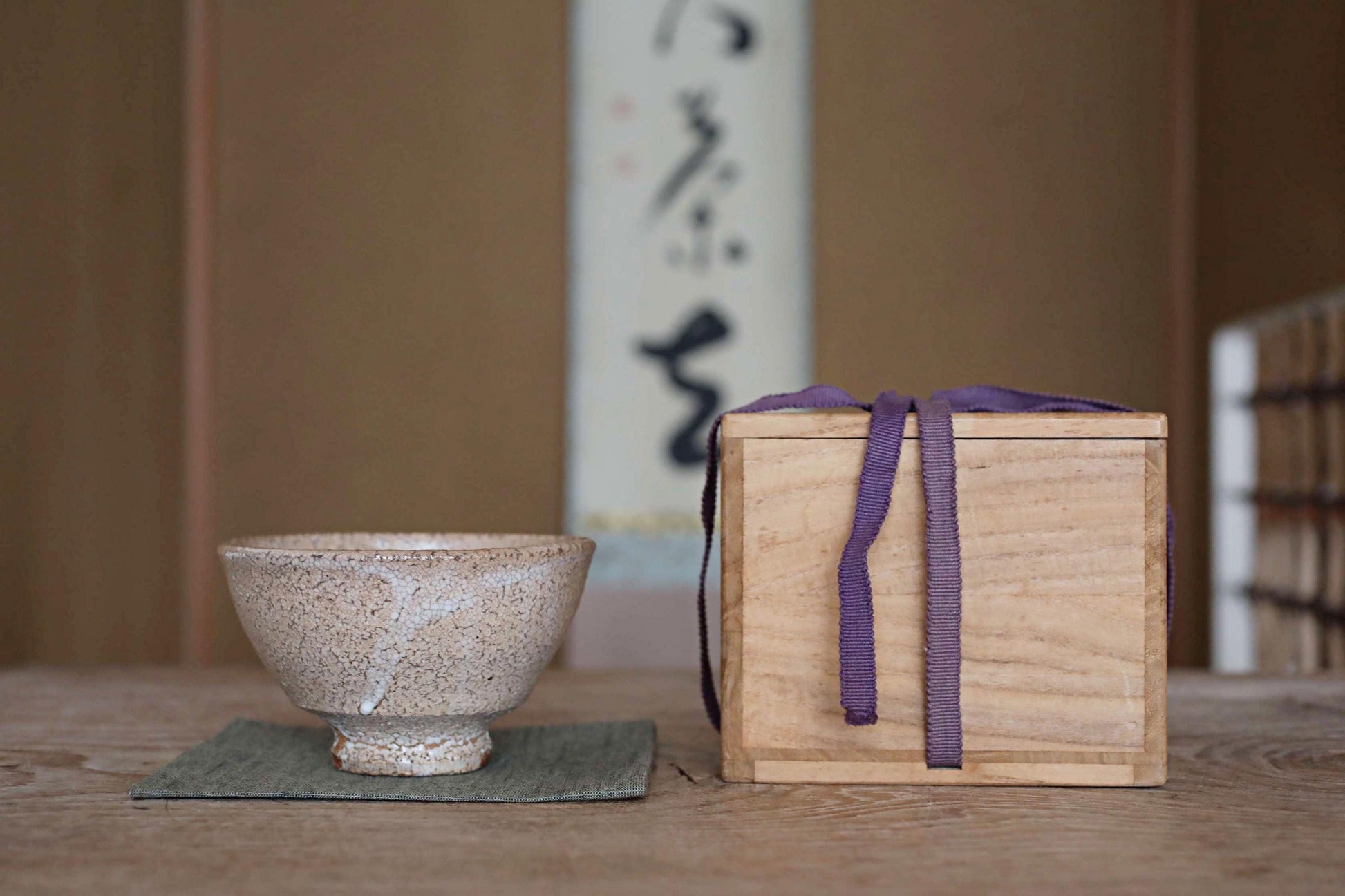 A white ceramic bowl and a wooden box with a purple ribbon are placed on a wooden table, with a white wall and a black and white Japanese calligraphy poster in the background.