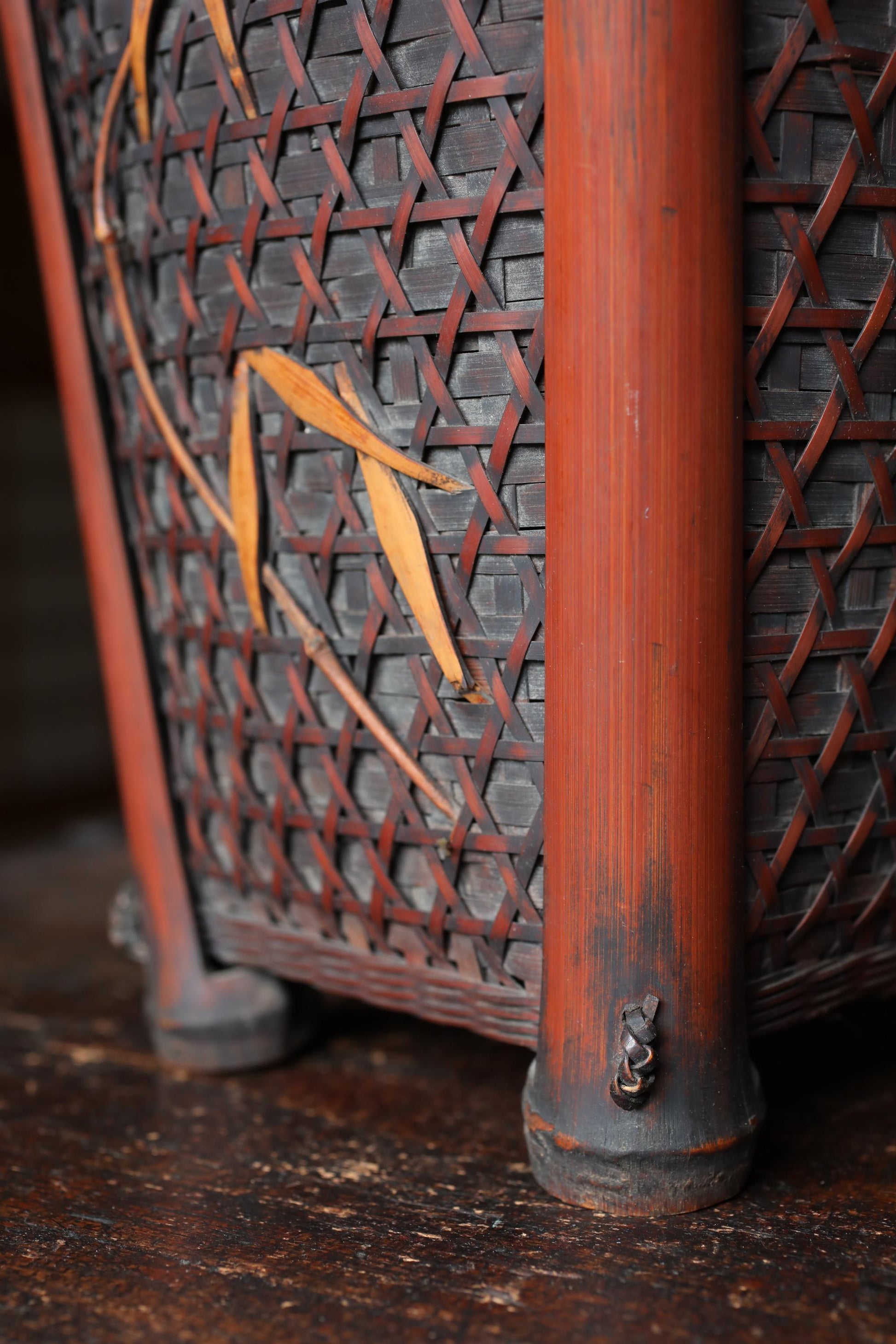 a close-up view of a woven basket with a reddish-brown color and a pattern of interwoven bamboo strips. The basket appears to be made of wood and has a metal clasp on the bottom.