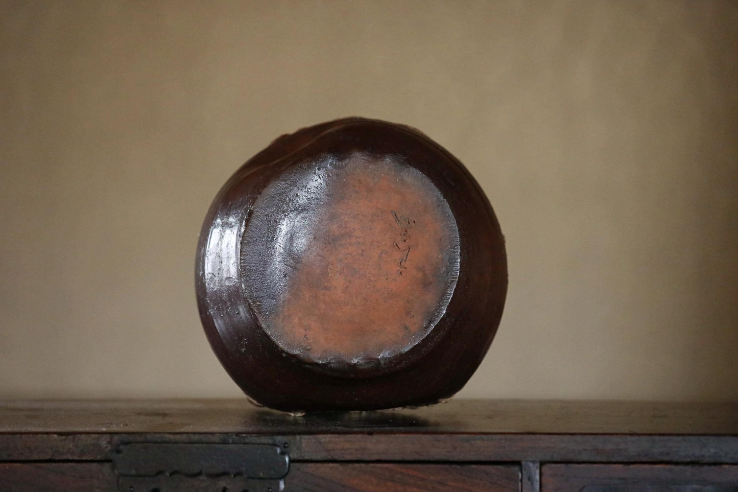A dark brown, round ceramic vase or container sits on a wooden surface against a beige wall.