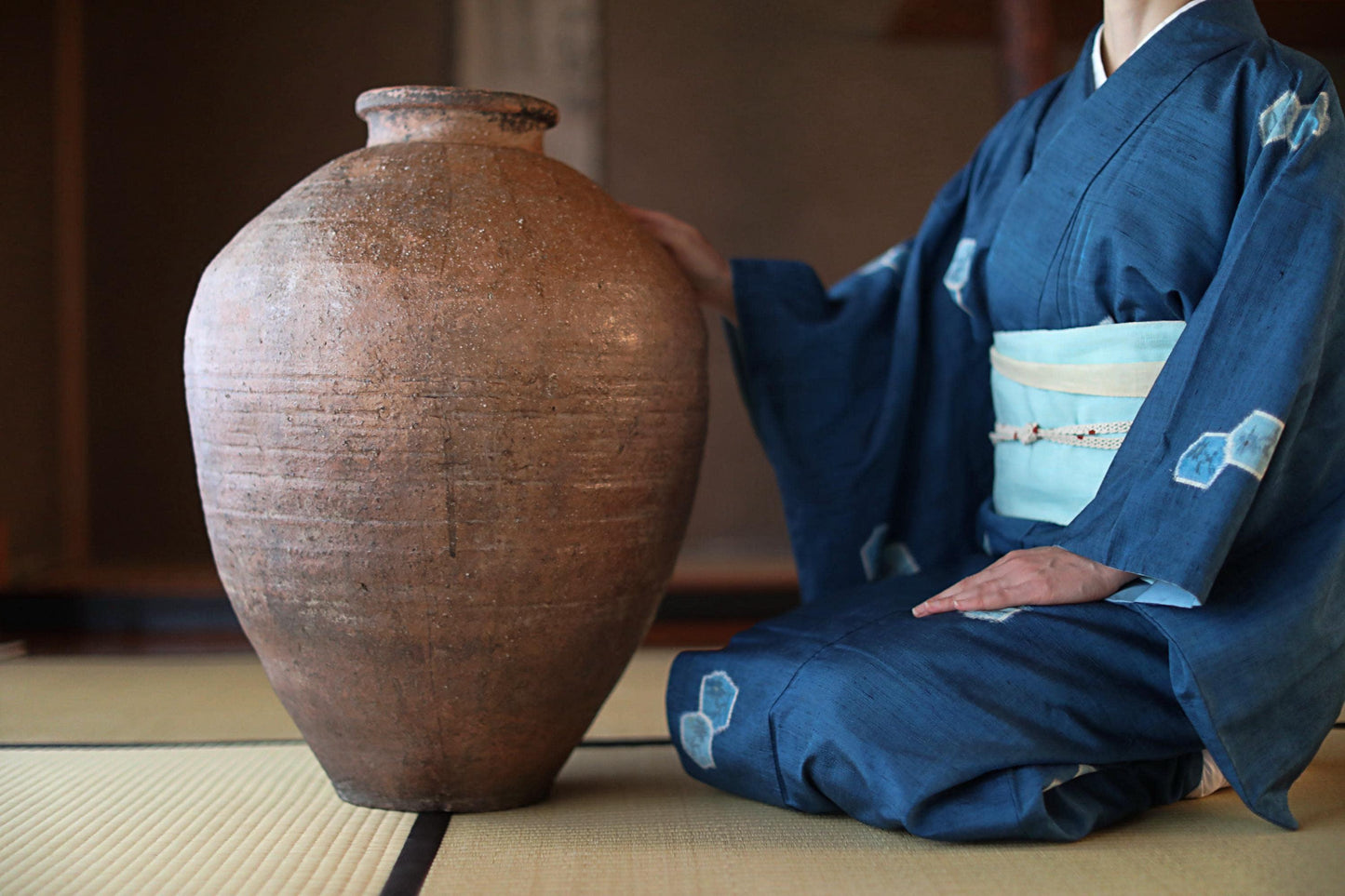 A person in a blue kimono is sitting on the floor next to a large, brown vase.
