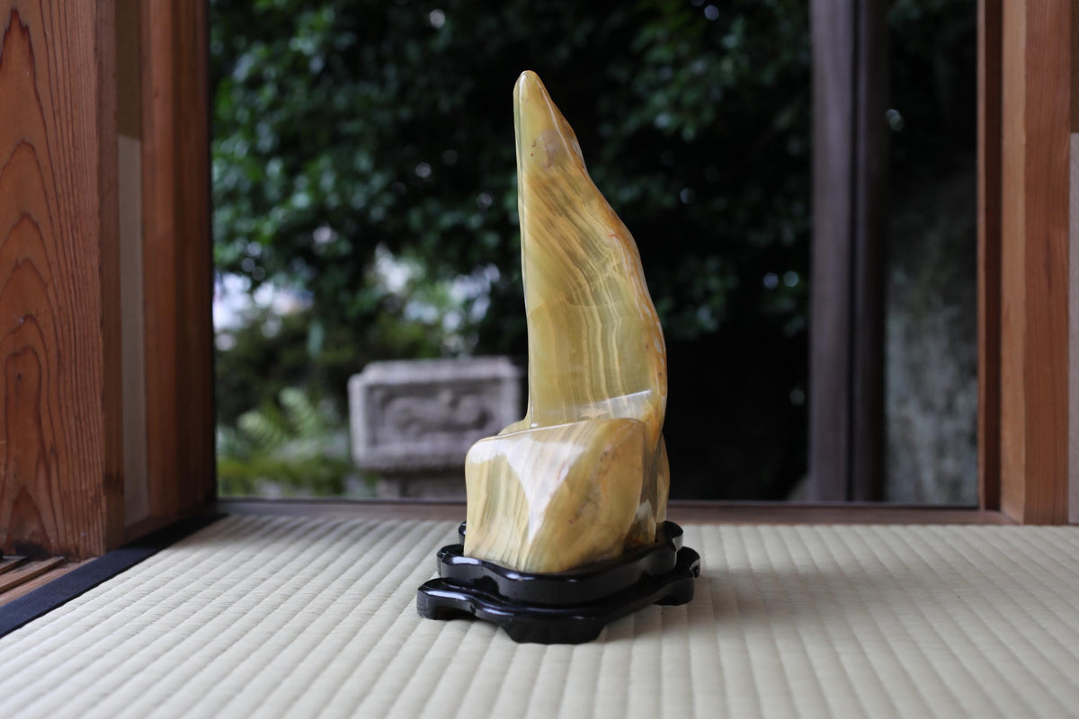 A yellow, curved stone sculpture is displayed on a black stand in a room with a wooden frame.