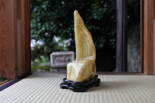 A yellow, curved stone sculpture is displayed on a black stand in a room with a wooden frame.