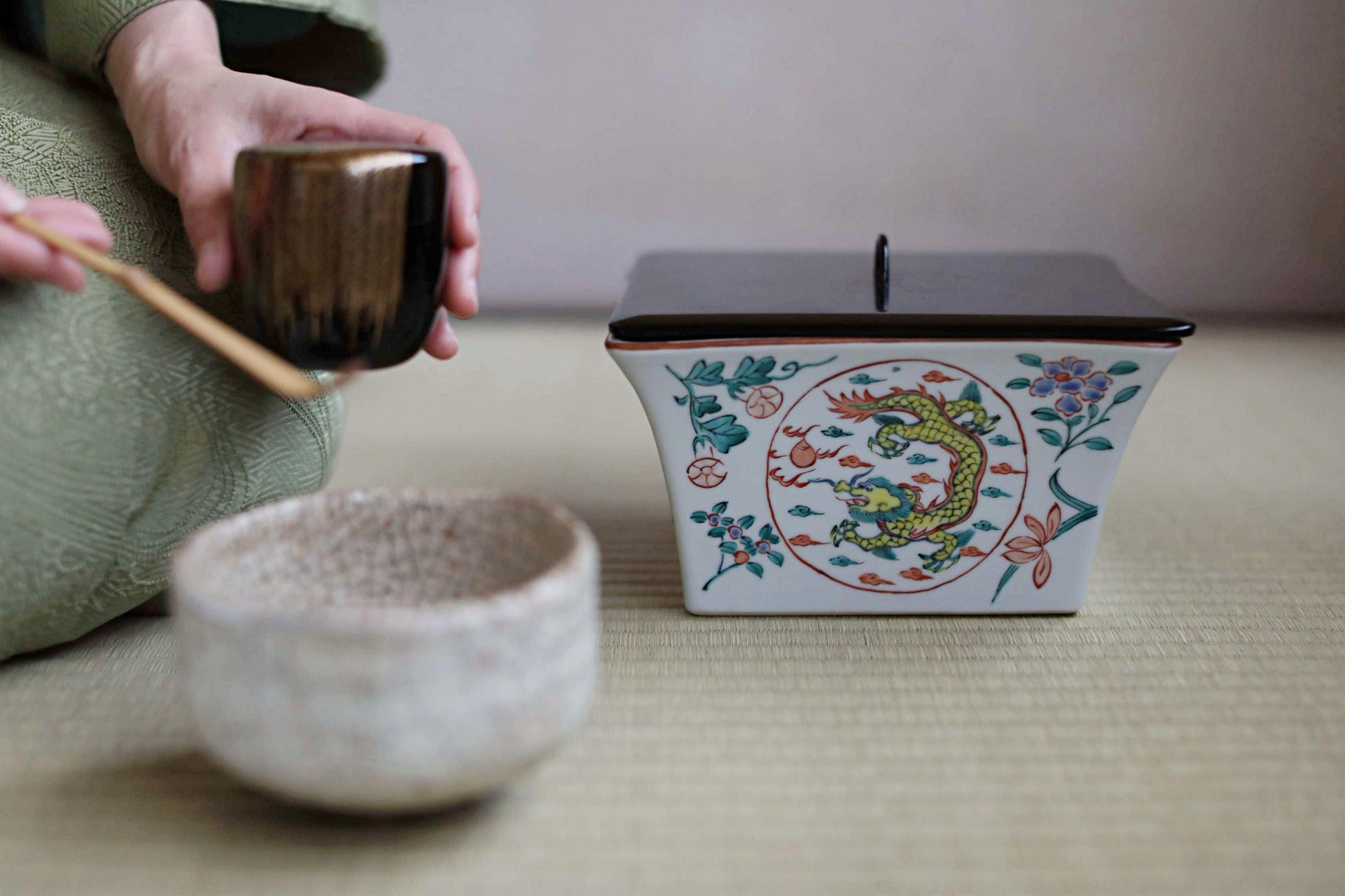 A person is sitting on the floor, holding a small bowl and a small cup, with a decorative ceramic container featuring a dragon design nearby.