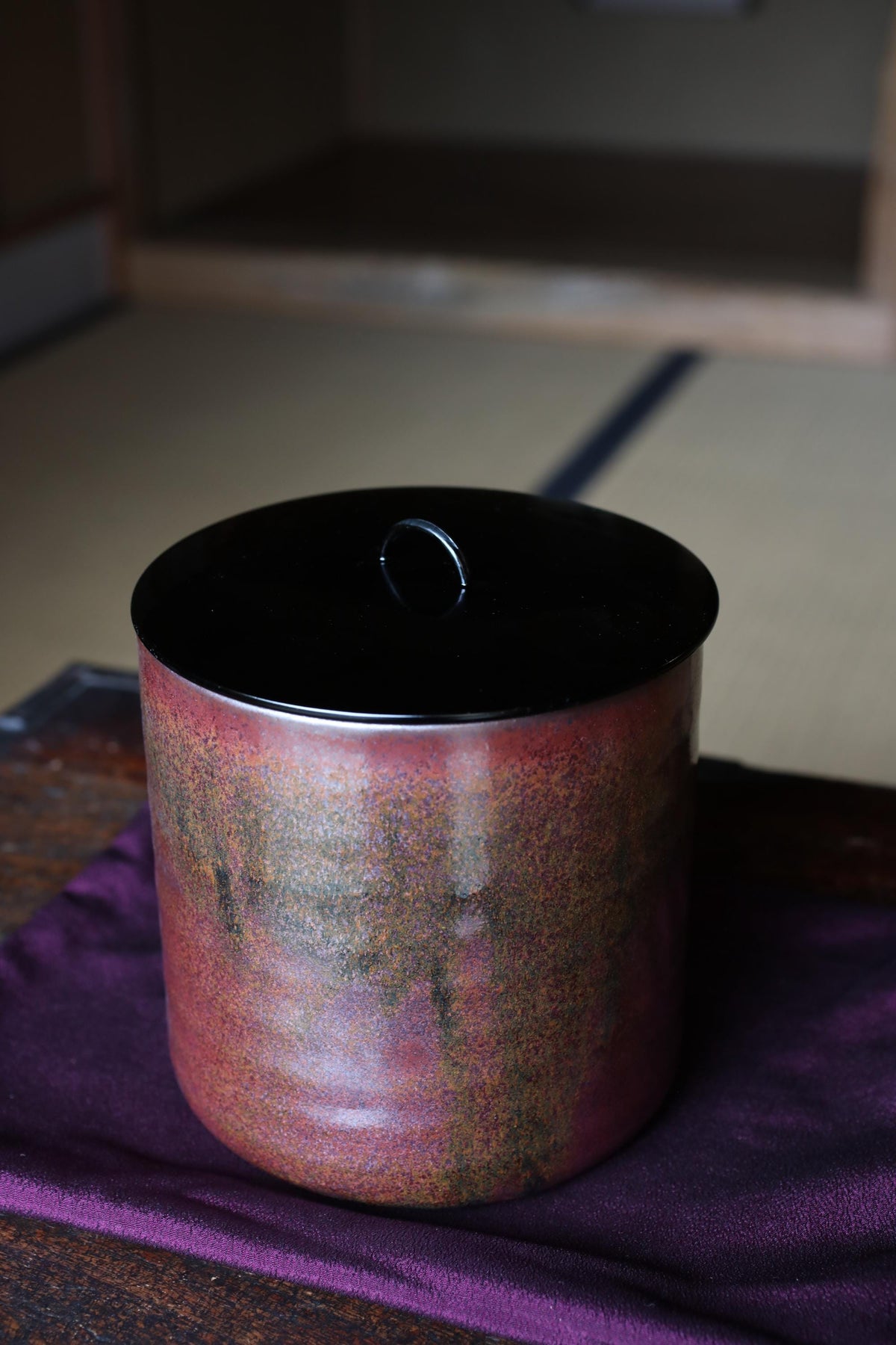 A ceramic container with a black lid sits on a purple cloth, with a blurred background suggesting a room setting.