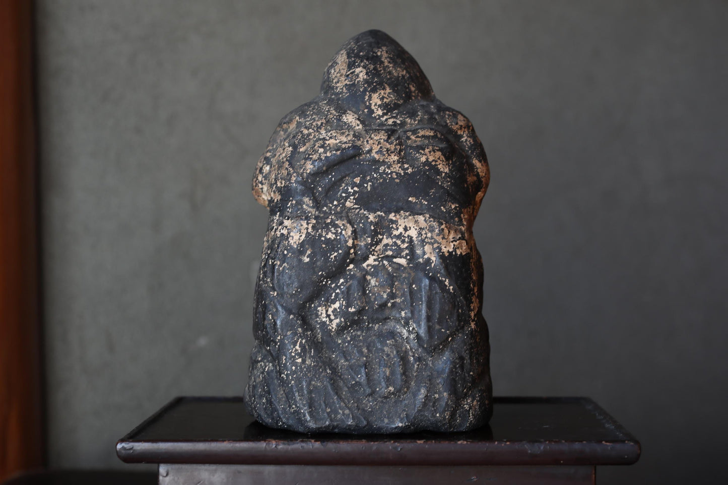 a black, weathered stone statue of a human figure, sitting on a wooden pedestal.