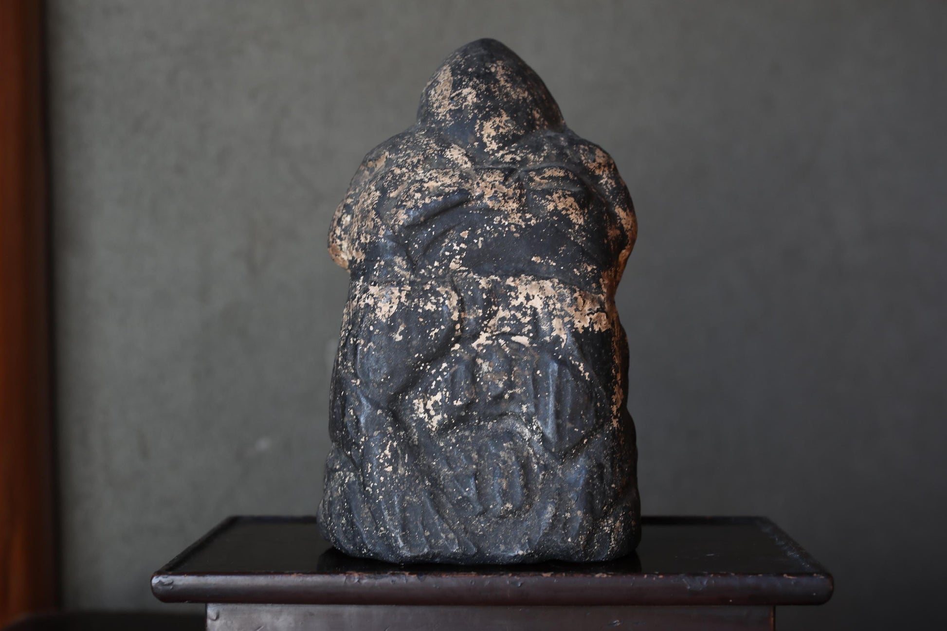 a black, weathered stone statue of a human figure, sitting on a wooden pedestal.