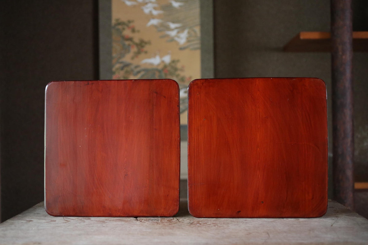 Two square wooden planks are placed side by side on a wooden surface, with a blurred background that includes a wall and a window.