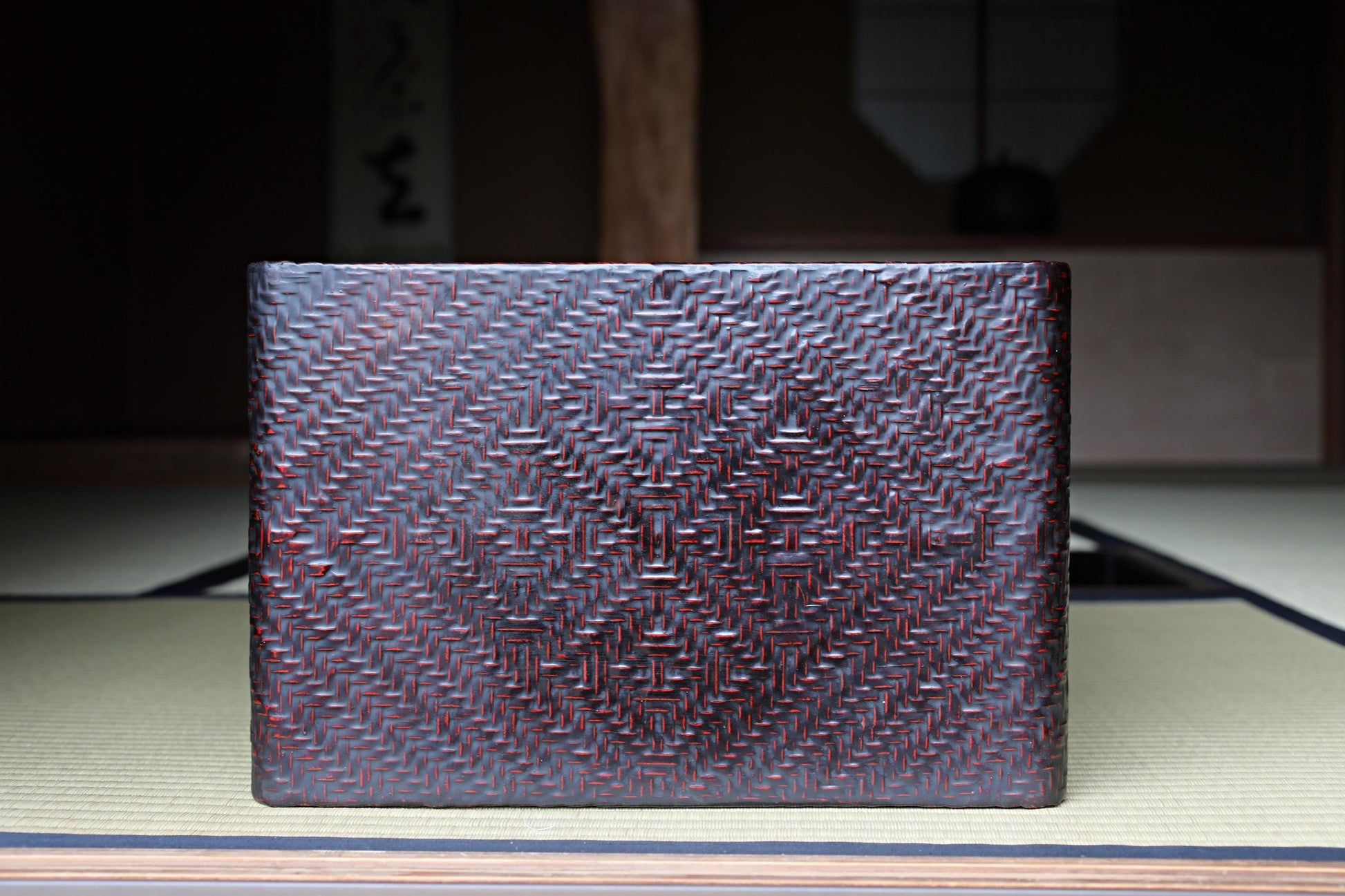 A dark, textured woven object with a geometric pattern, possibly a piece of furniture or a decorative item, placed on a wooden surface.