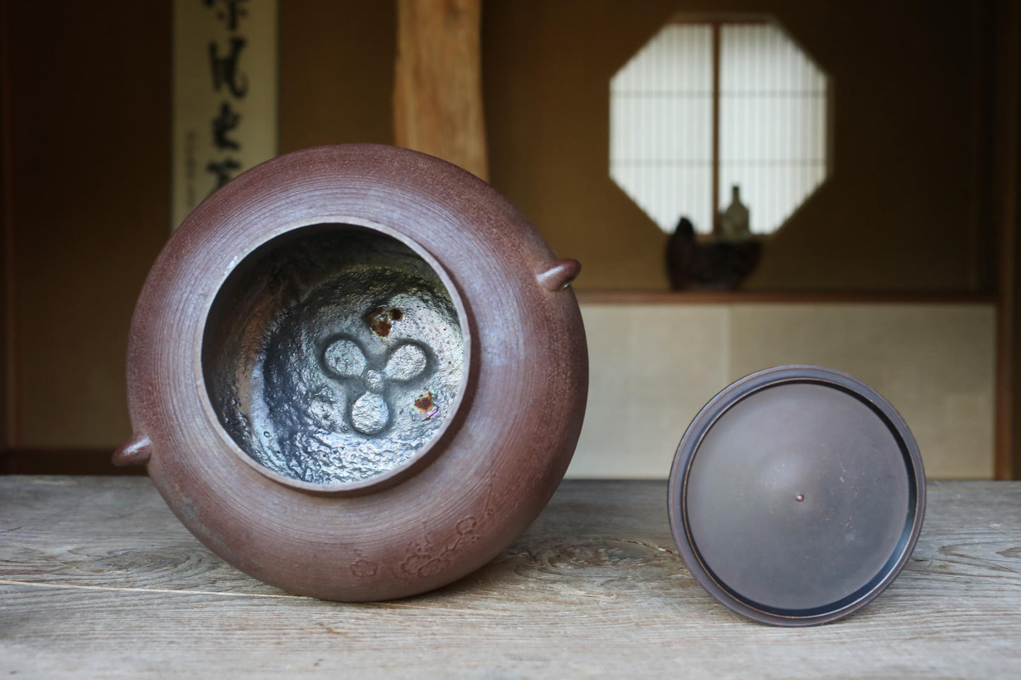 A brown ceramic tea pot with a lid sits on a wooden surface, with a window and a decorative object visible in the background.