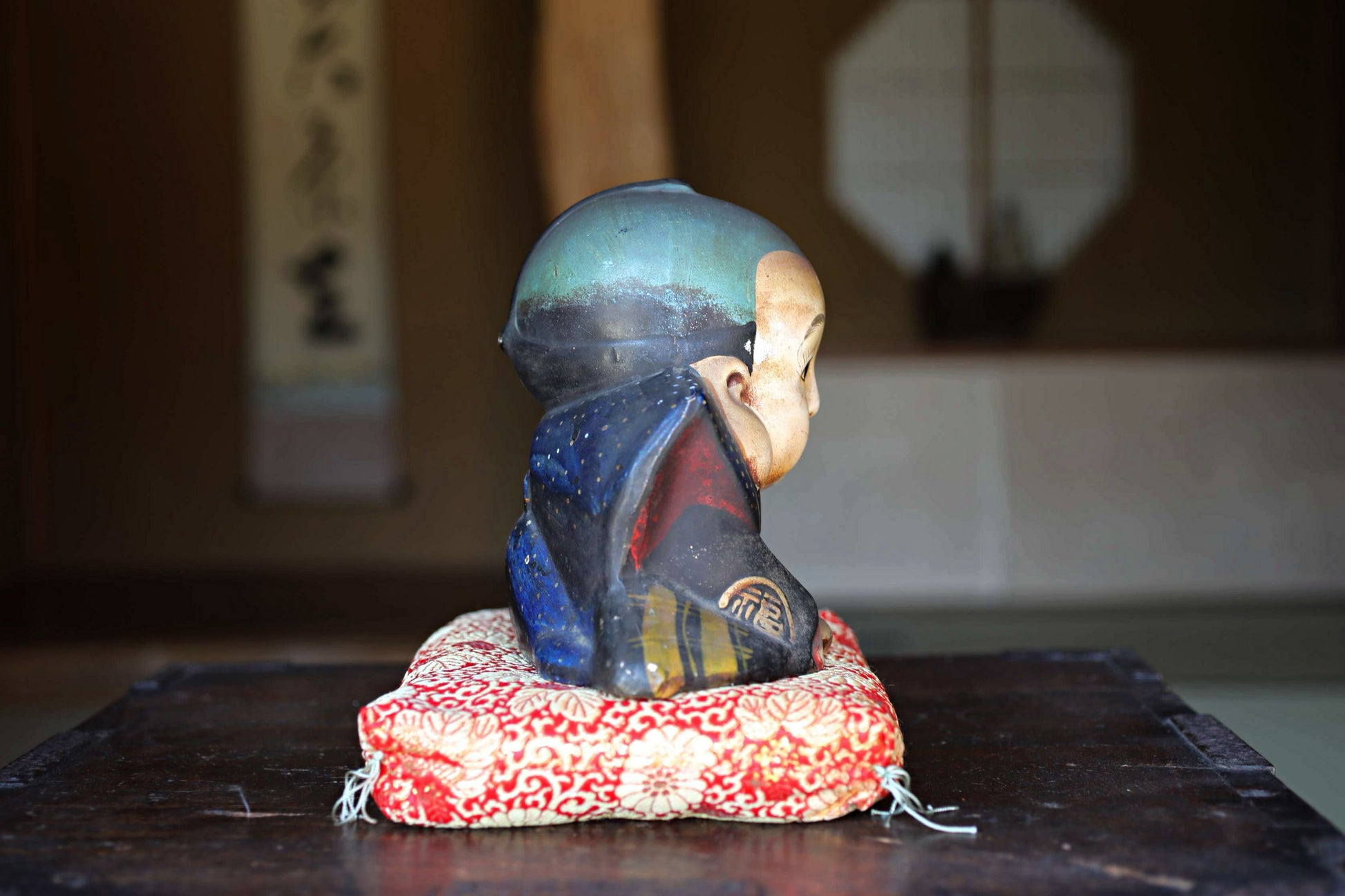 A ceramic figurine of a person sitting on a cushion, placed on a wooden surface.