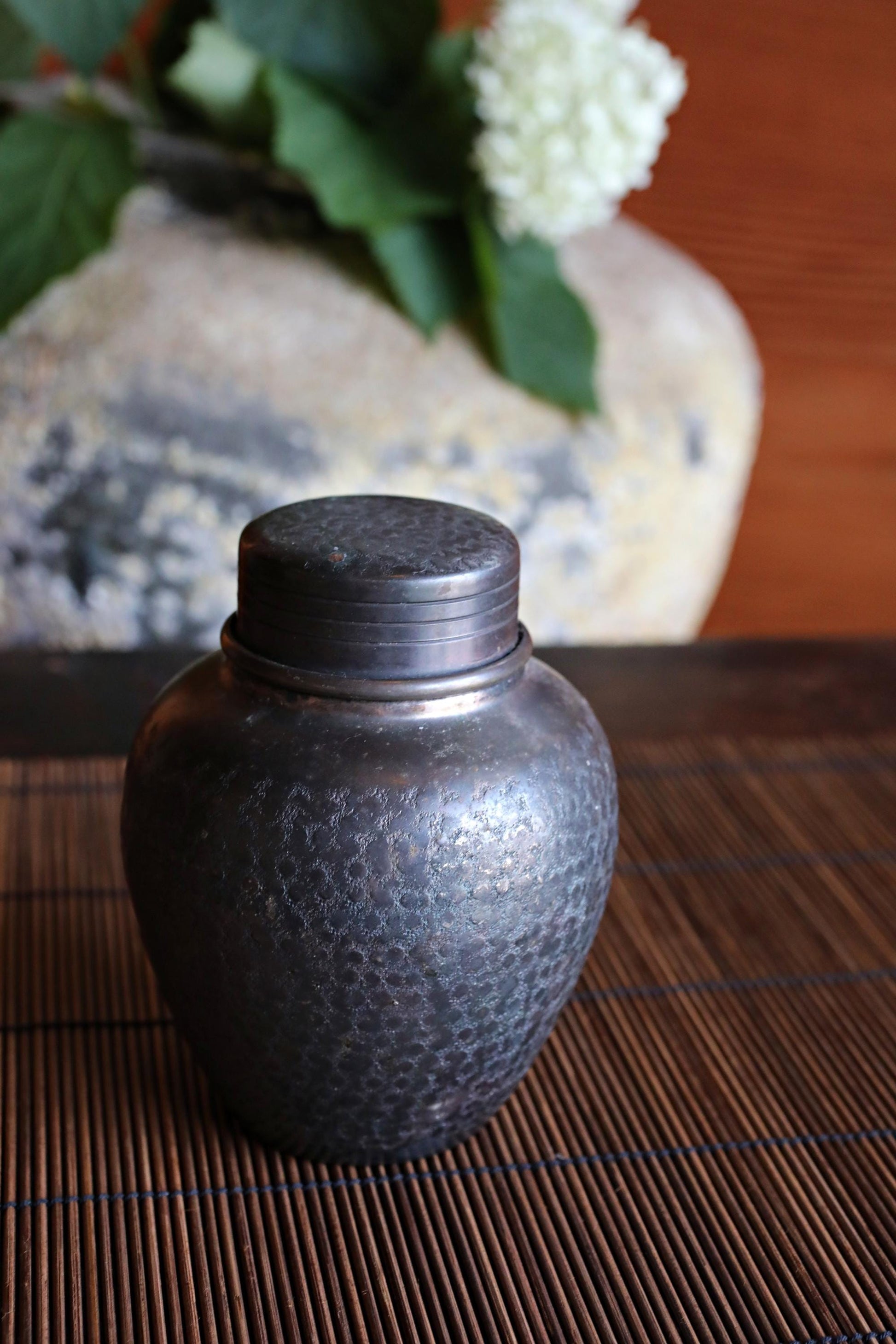 A dark metal jar with a lid sits on a bamboo mat, with a vase of white flowers in the background.
