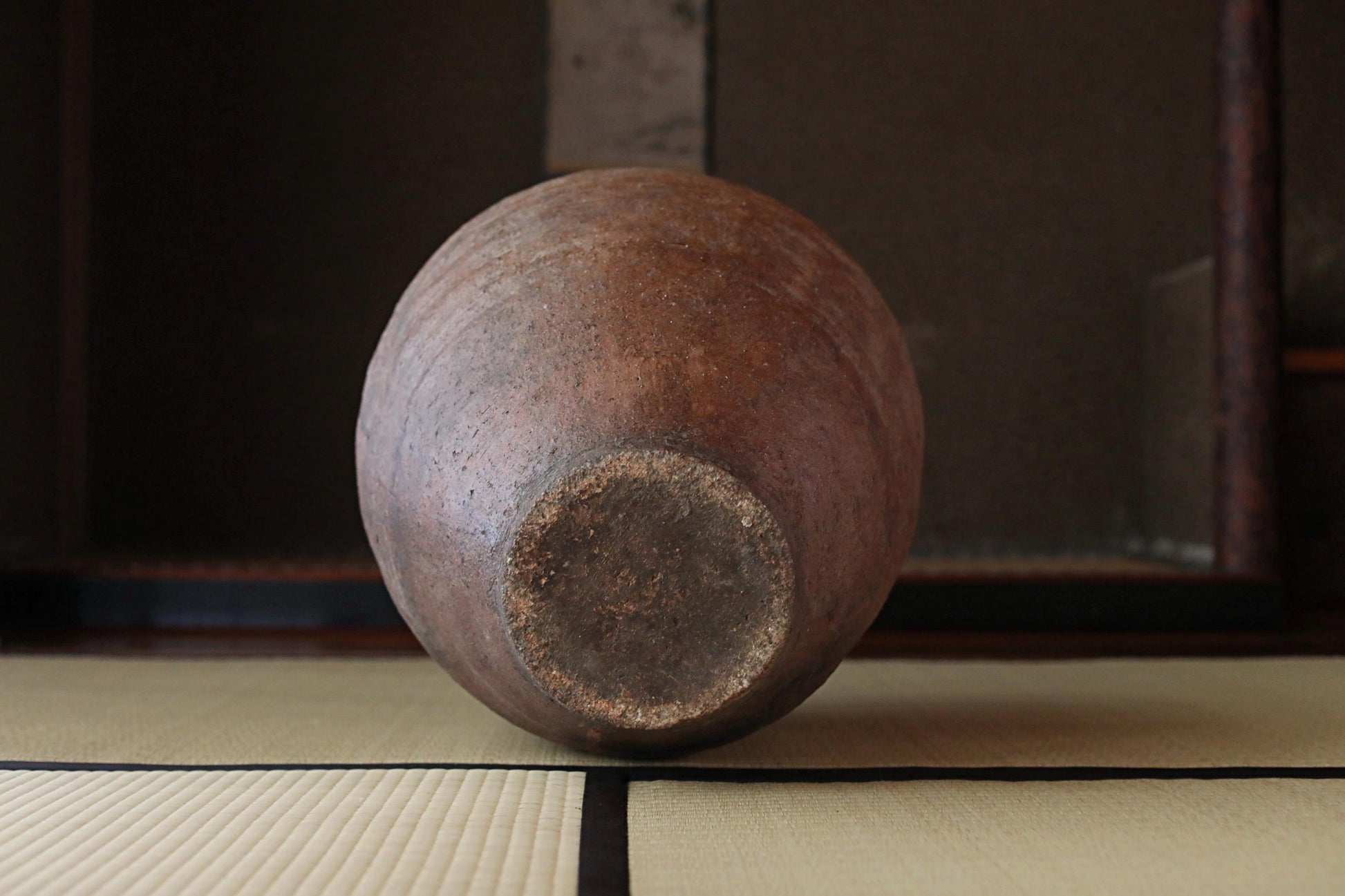 An old, round clay pot sits on a tatami mat, its weathered surface hinting at its age and history.