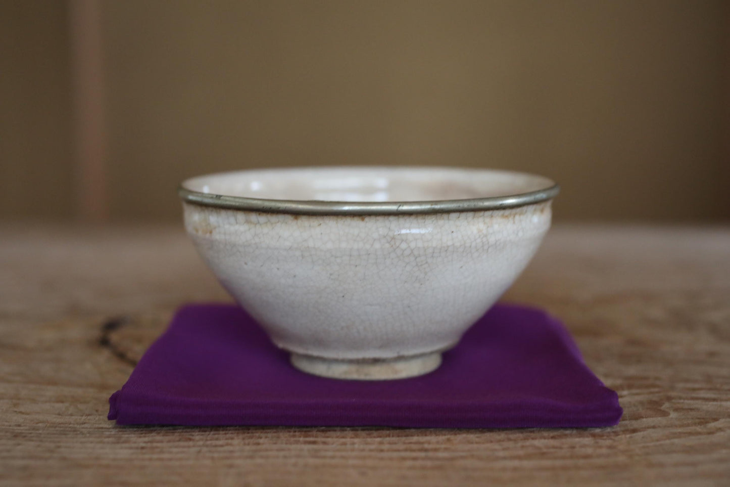 A white ceramic bowl with a silver rim sits on a purple cloth on a wooden surface.