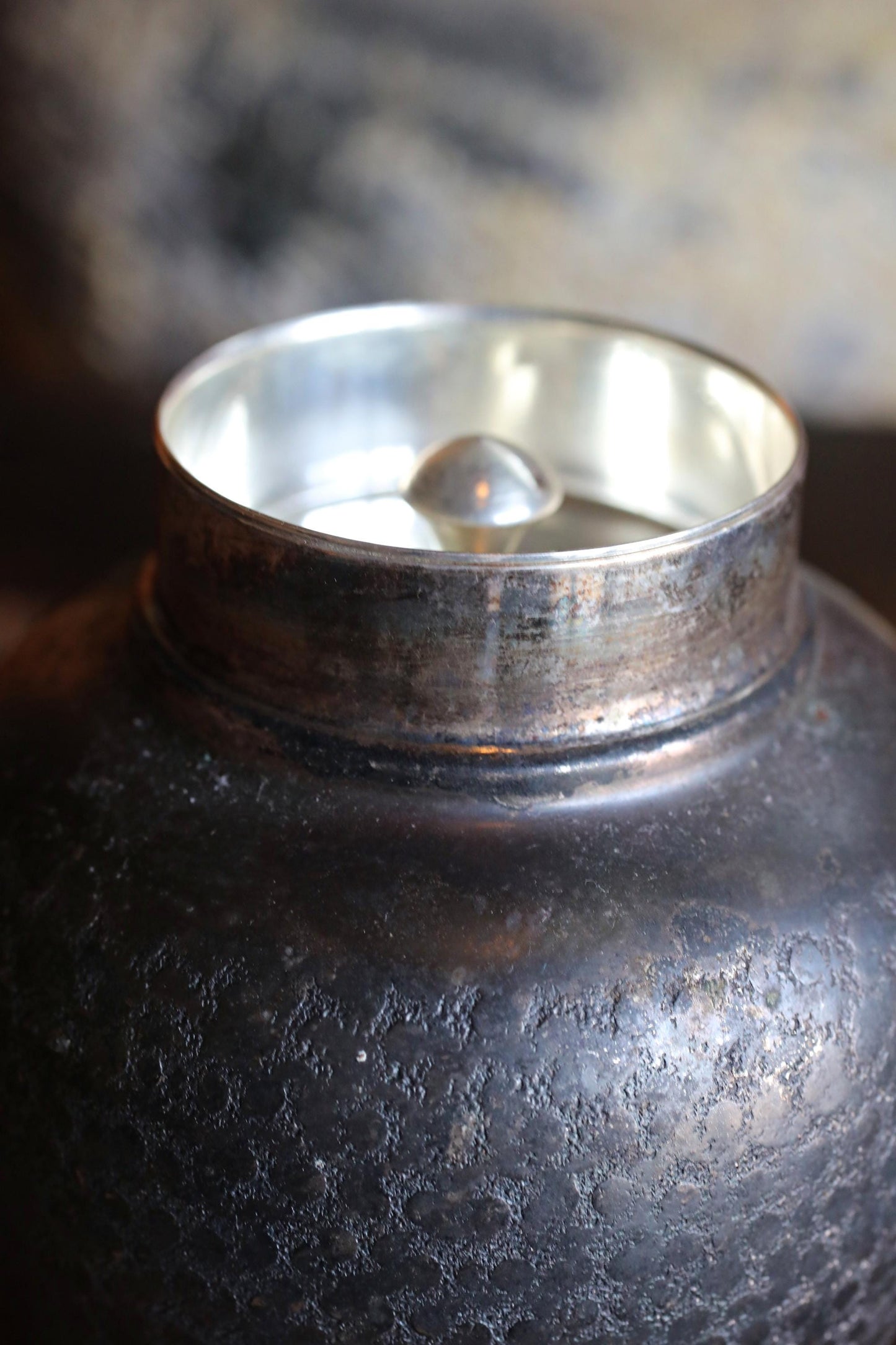 A close-up view of a dark, textured metal object with a silver-colored lid or cover, possibly a container or jar.