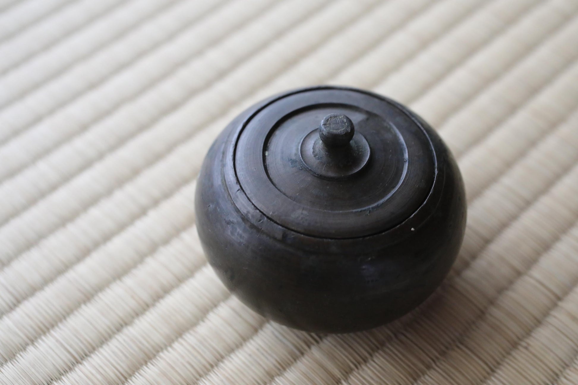 A black, round ceramic container with a lid sits on a woven mat.