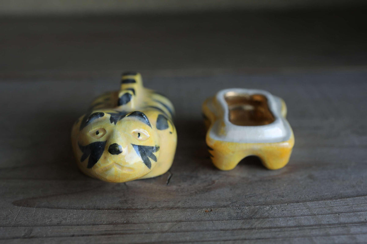 A ceramic figurine of a tiger-like animal, possibly a cat or a tiger, is sitting on a wooden surface next to an open ceramic container.