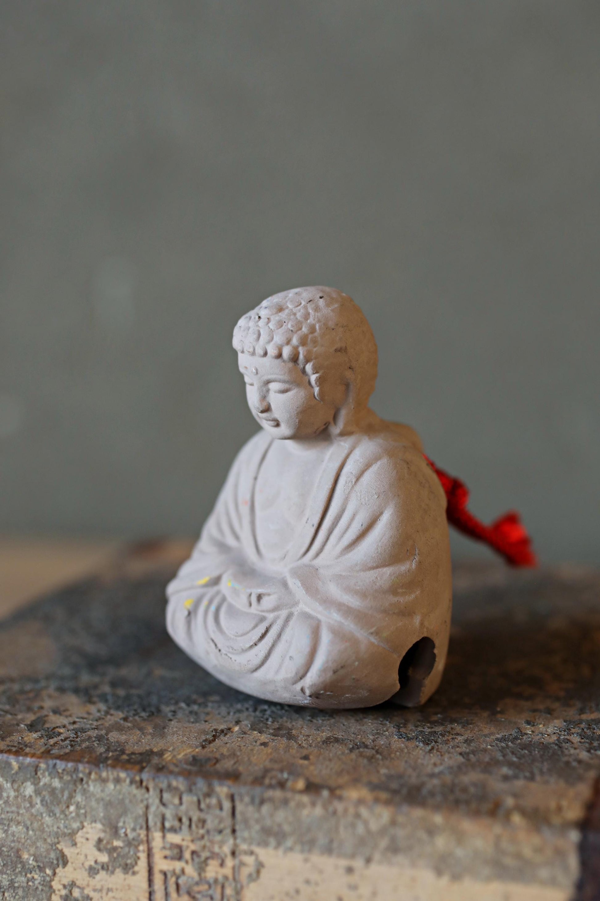 A small white Buddha statue sitting on a gray surface, with a red ribbon or cloth draped around its neck.