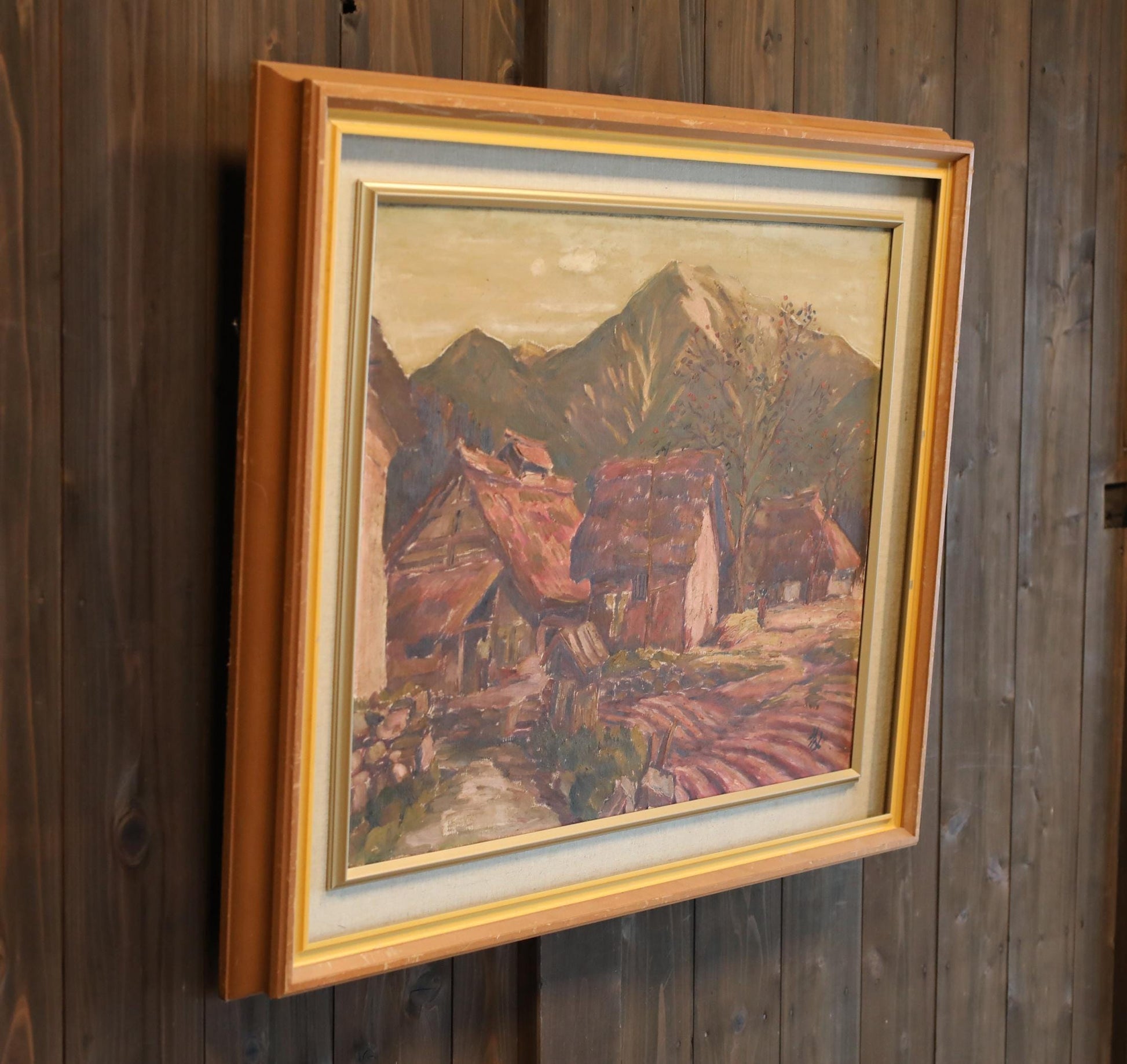 a framed painting of a rural landscape with mountains in the background.