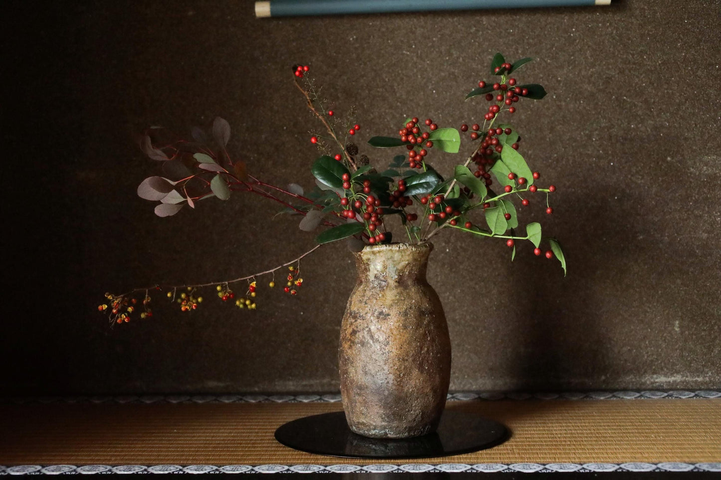 A rustic vase filled with a mix of red berries and green leaves sits on a wooden surface against a dark background.