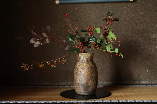 A rustic vase filled with a mix of red berries and green leaves sits on a wooden surface against a dark background.