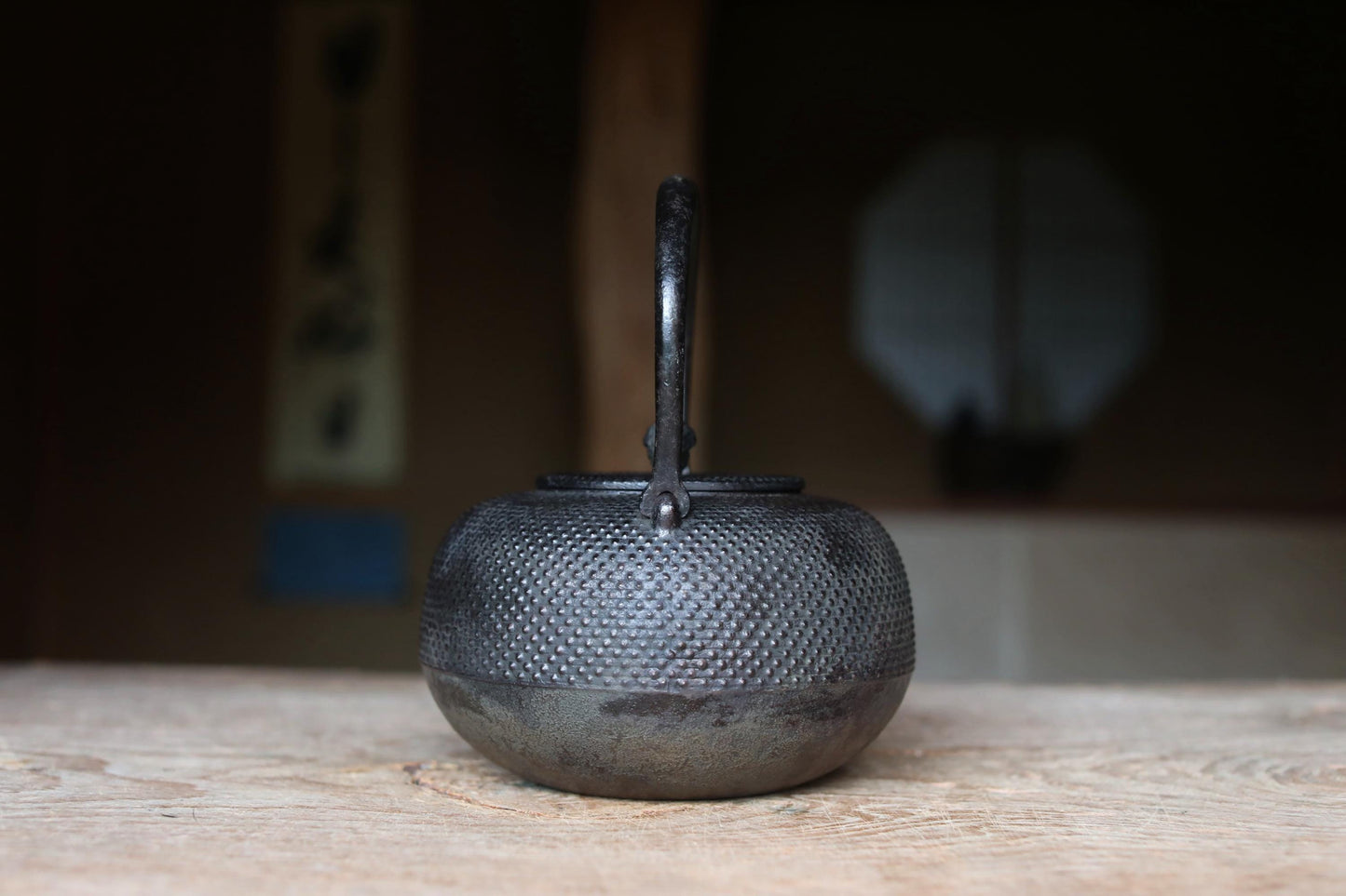 A black metal teapot with a textured surface sits on a wooden surface, with a blurred background suggesting an indoor setting.