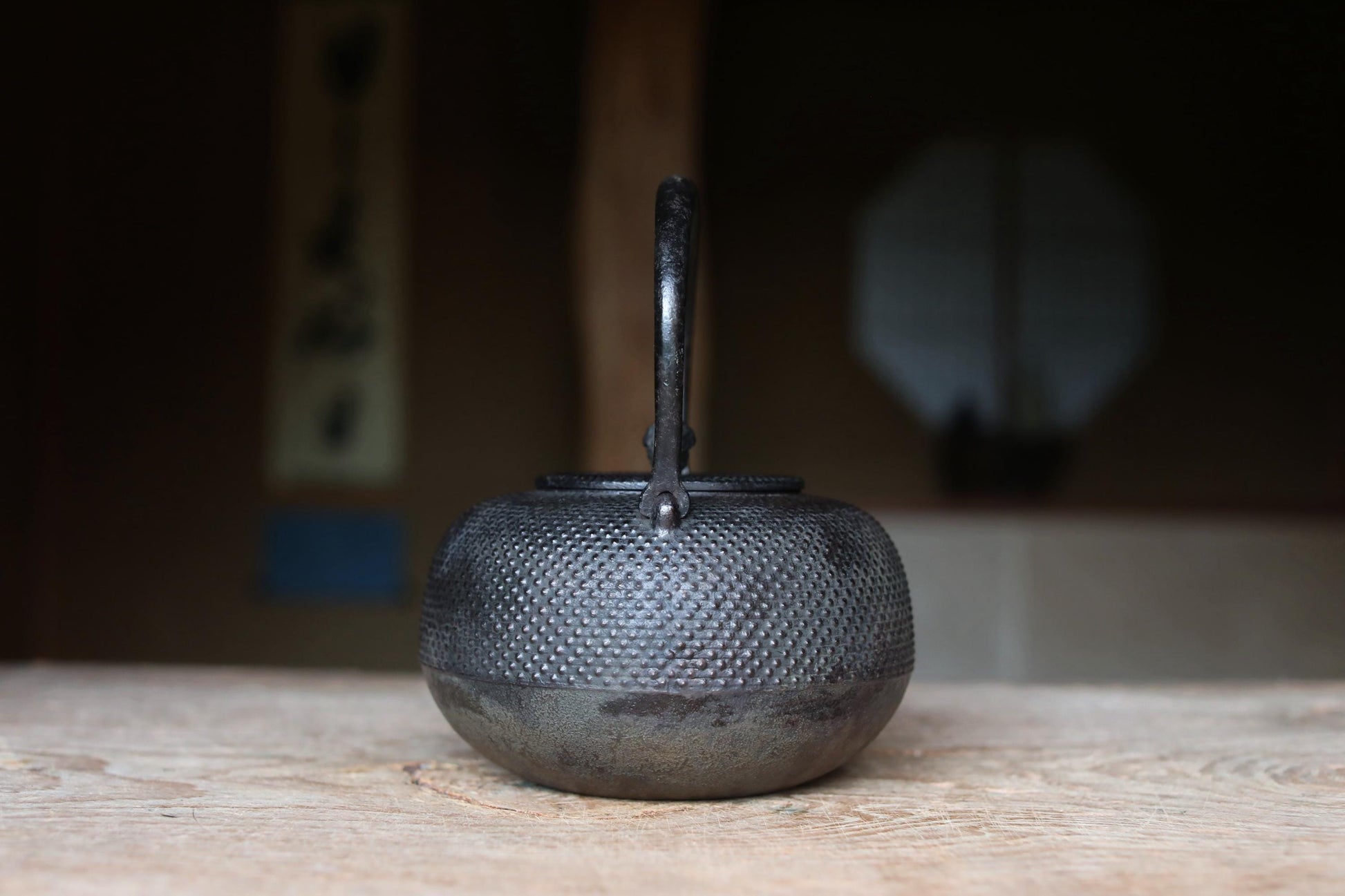 A black metal teapot with a textured surface sits on a wooden surface, with a blurred background suggesting an indoor setting.