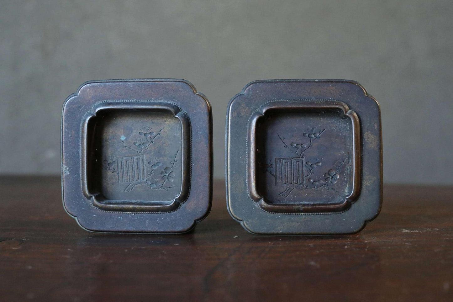 Two antique metal trays with a square design and a floral motif, placed side by side on a wooden surface.