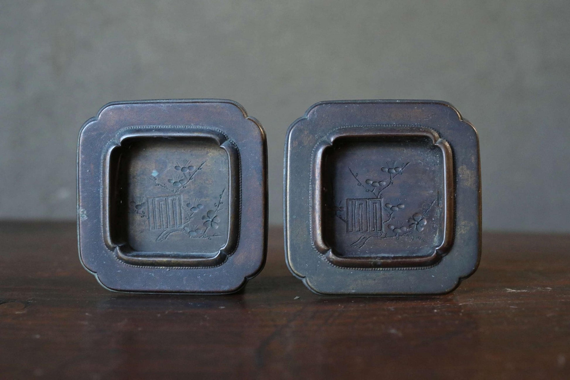 Two antique metal trays with a square design and a floral motif, placed side by side on a wooden surface.