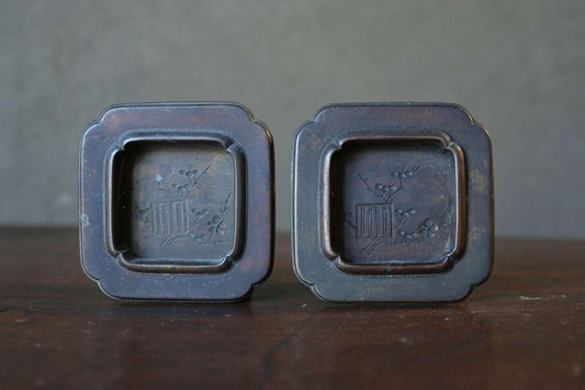 Two antique metal trays with a square design and a floral motif, placed side by side on a wooden surface.
