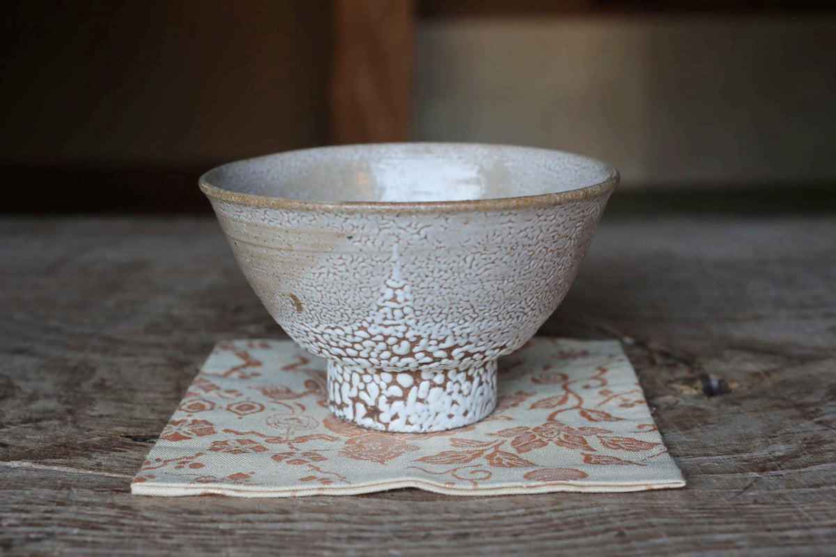 A white ceramic bowl with a speckled pattern sits on a beige napkin with a floral pattern on a wooden surface.
