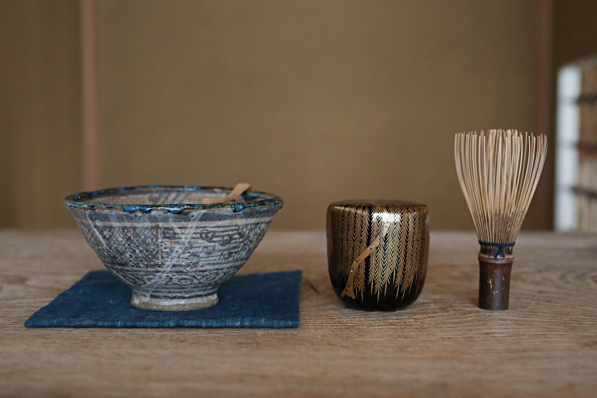 A blue and white ceramic bowl, a black and gold ceramic cup, and a wooden whisk are arranged on a wooden surface.