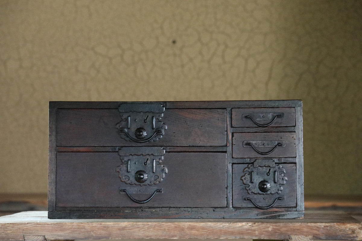 A dark wooden chest with multiple drawers and ornate metal handles is displayed on a wooden surface against a textured wall.