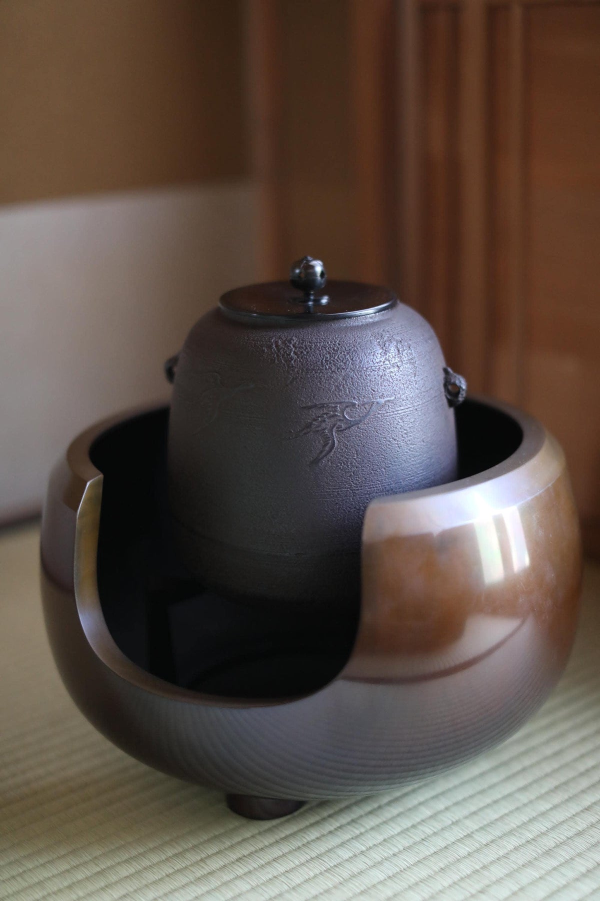 A dark-colored ceramic teapot with a lid sits on a wooden surface, surrounded by a wooden cabinet.