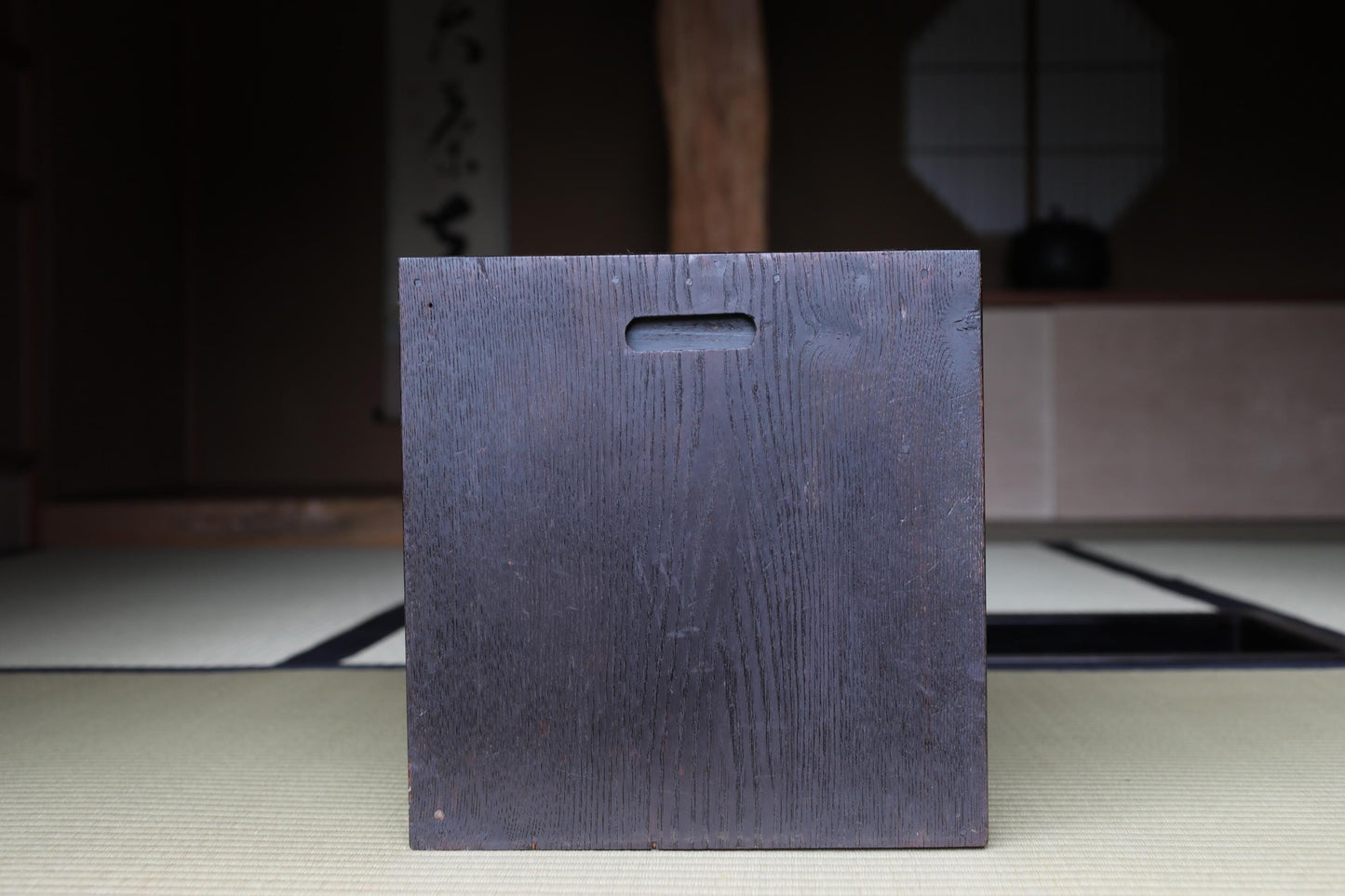 A dark wooden box with a hole in the center, placed on a tatami mat in a dimly lit room with a wooden beam and a window in the background.