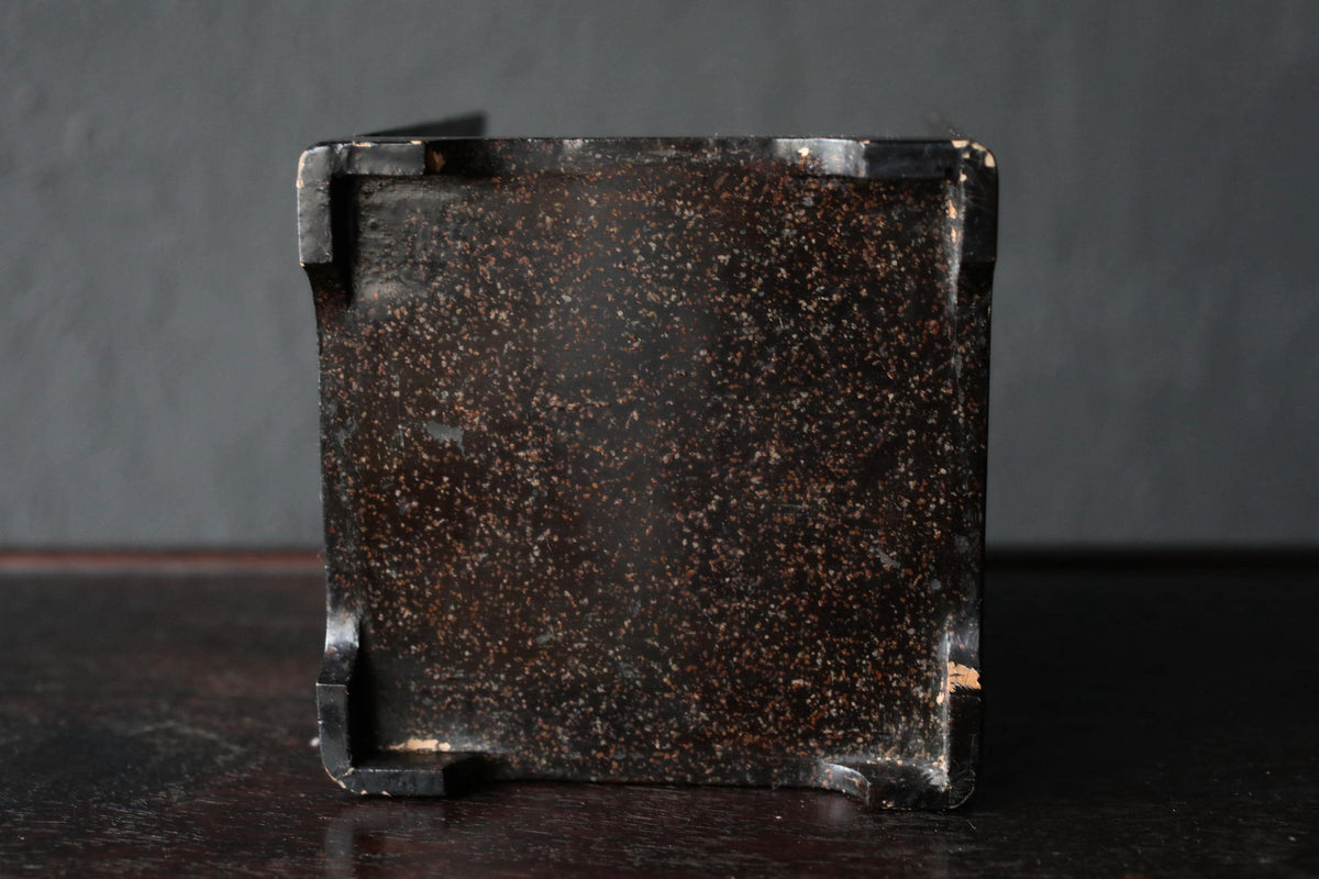 a square, dark-colored metal object with a speckled, rust-colored surface, sitting on a dark surface.