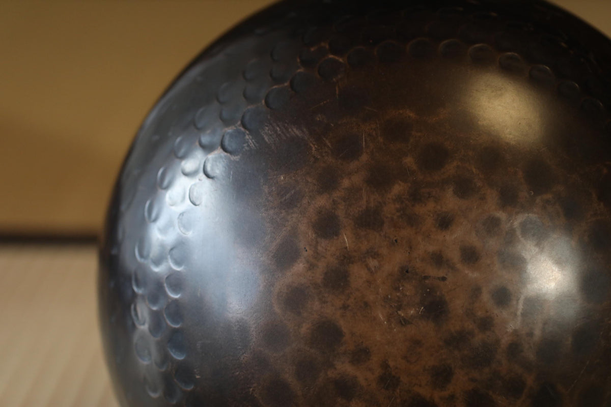 A close-up image of a dark, textured surface with a pattern of small circles or dots, possibly a metal or ceramic object.