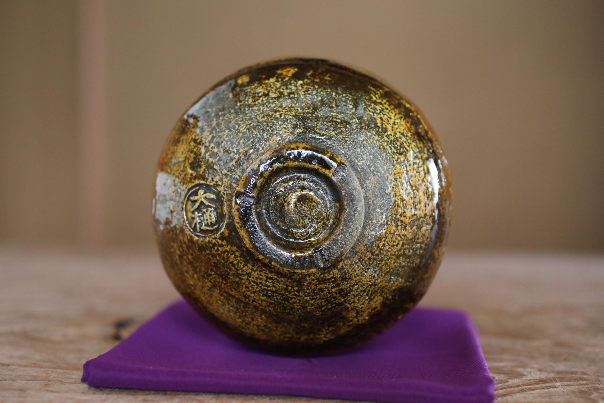 A close-up image of a round, golden-colored ceramic object, possibly a vase or bowl, placed on a purple cloth or cushion.