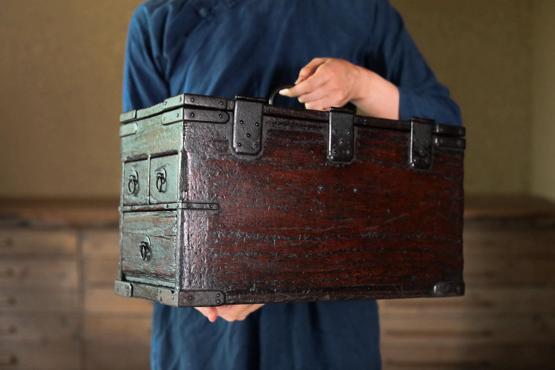 A person wearing a blue shirt is holding a large, old-fashioned wooden chest with metal clasps.