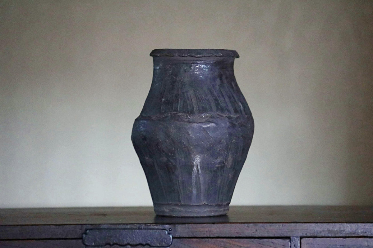A dark-colored vase with a textured surface sits on a wooden surface against a plain background.