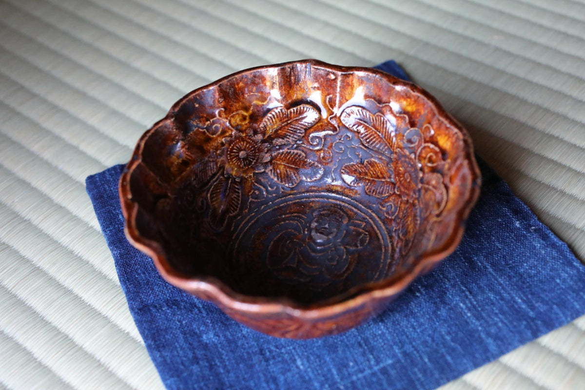 A brown ceramic bowl with intricate floral designs sits on a blue cloth.