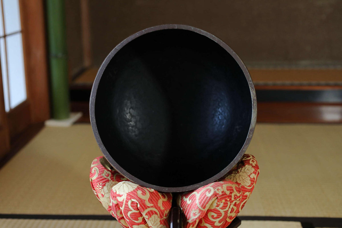 A black bowl is held in a pair of red and white patterned gloves, placed on a tatami mat in a room with a wooden floor.