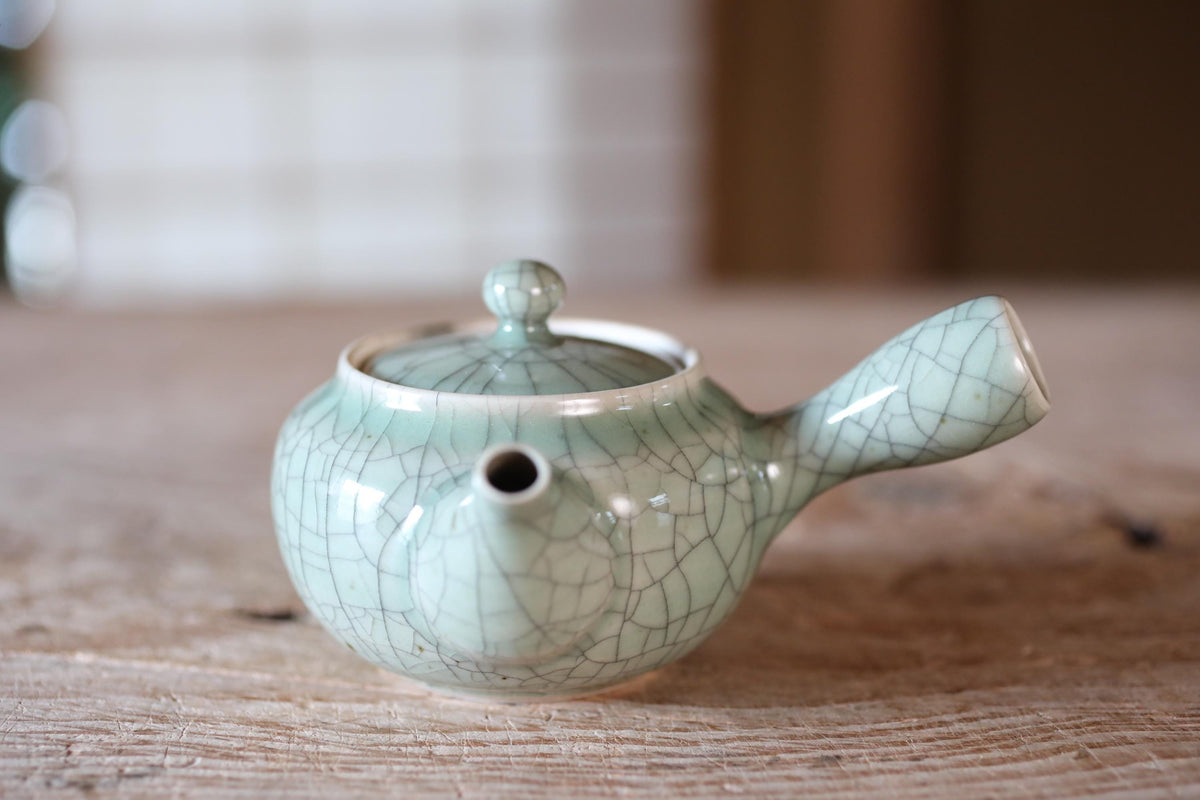 A ceramic teapot with a white and green color scheme, featuring a handle and spout, sits on a wooden surface.
