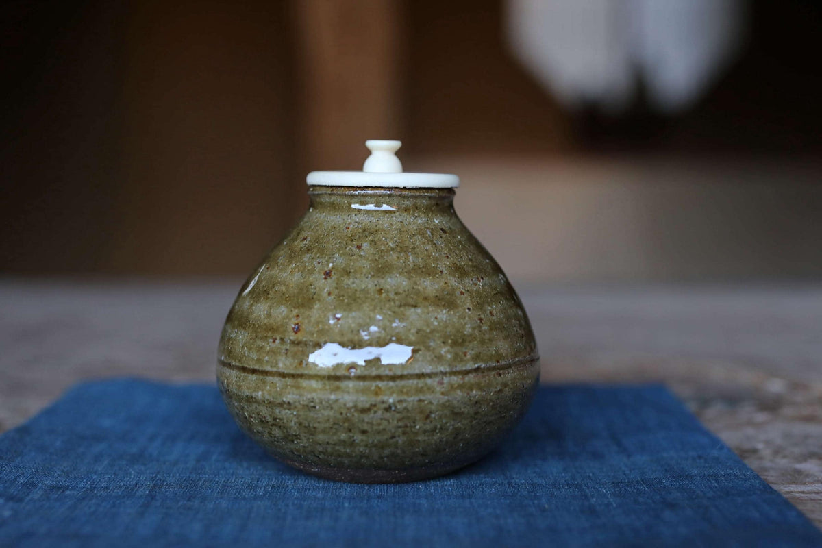 A ceramic jar with a white lid sits on a blue cloth, with a blurred background.