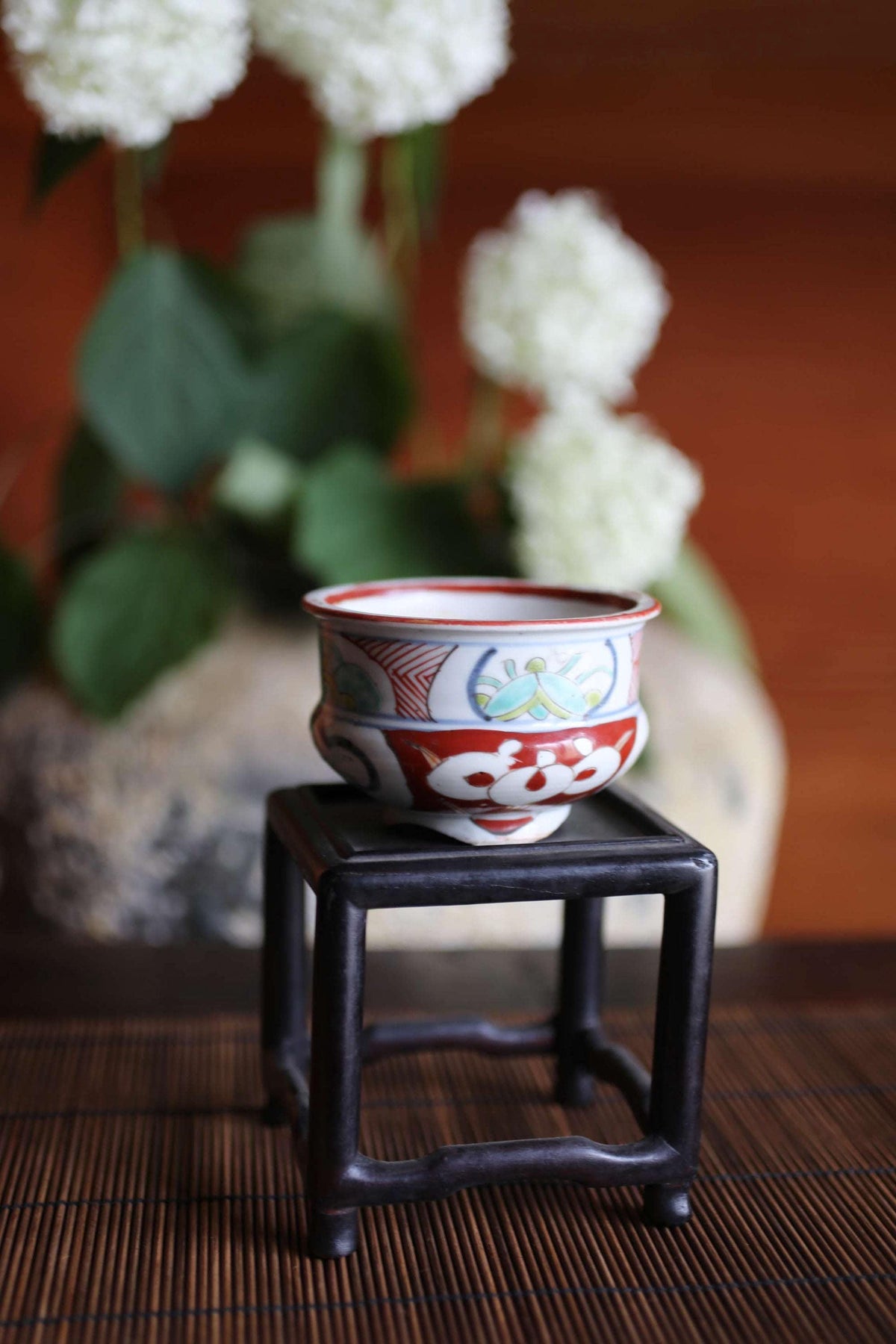 A decorative ceramic bowl with a floral pattern sits on a black stand, with white flowers in the background.