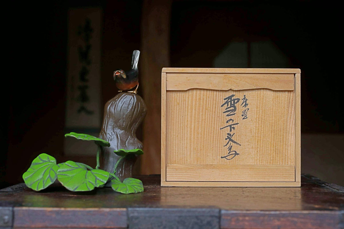A small figurine of a bird perched on a tree trunk, with a wooden box featuring Japanese characters next to it, both placed on a wooden surface.