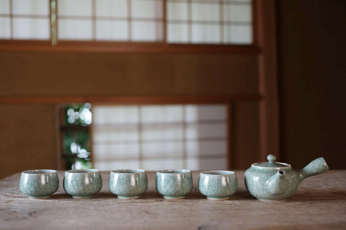 A row of six small, white ceramic cups arranged on a wooden surface, with a teapot and lid placed next to them.