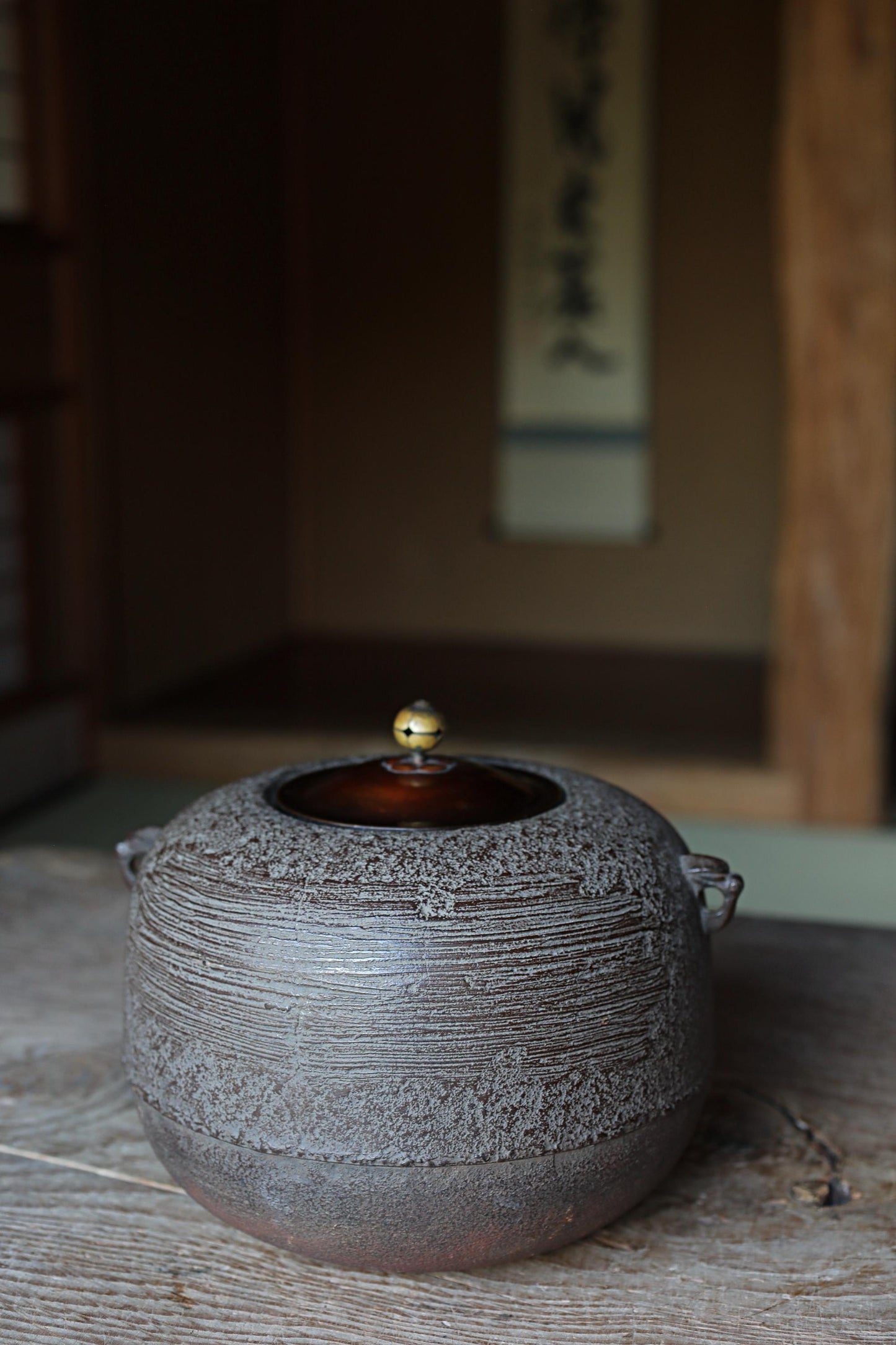 A ceramic teapot with a dark, textured surface sits on a wooden surface, with a blurred background that includes a wooden wall and a sign with Japanese characters.