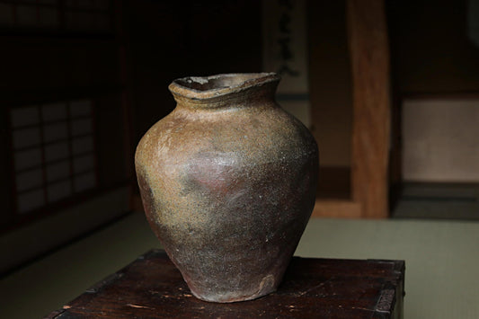 An old, weathered clay vase sits on a wooden surface, with a traditional Japanese-style room in the background.