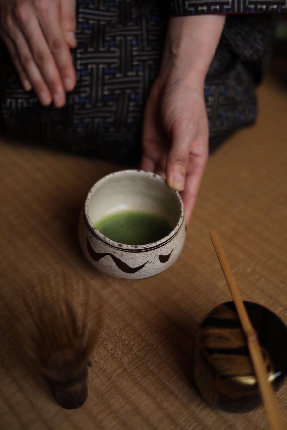 A person's hand is holding a small white bowl with a green liquid, likely tea, on a wooden surface. There is also a small brush and a small container nearby.