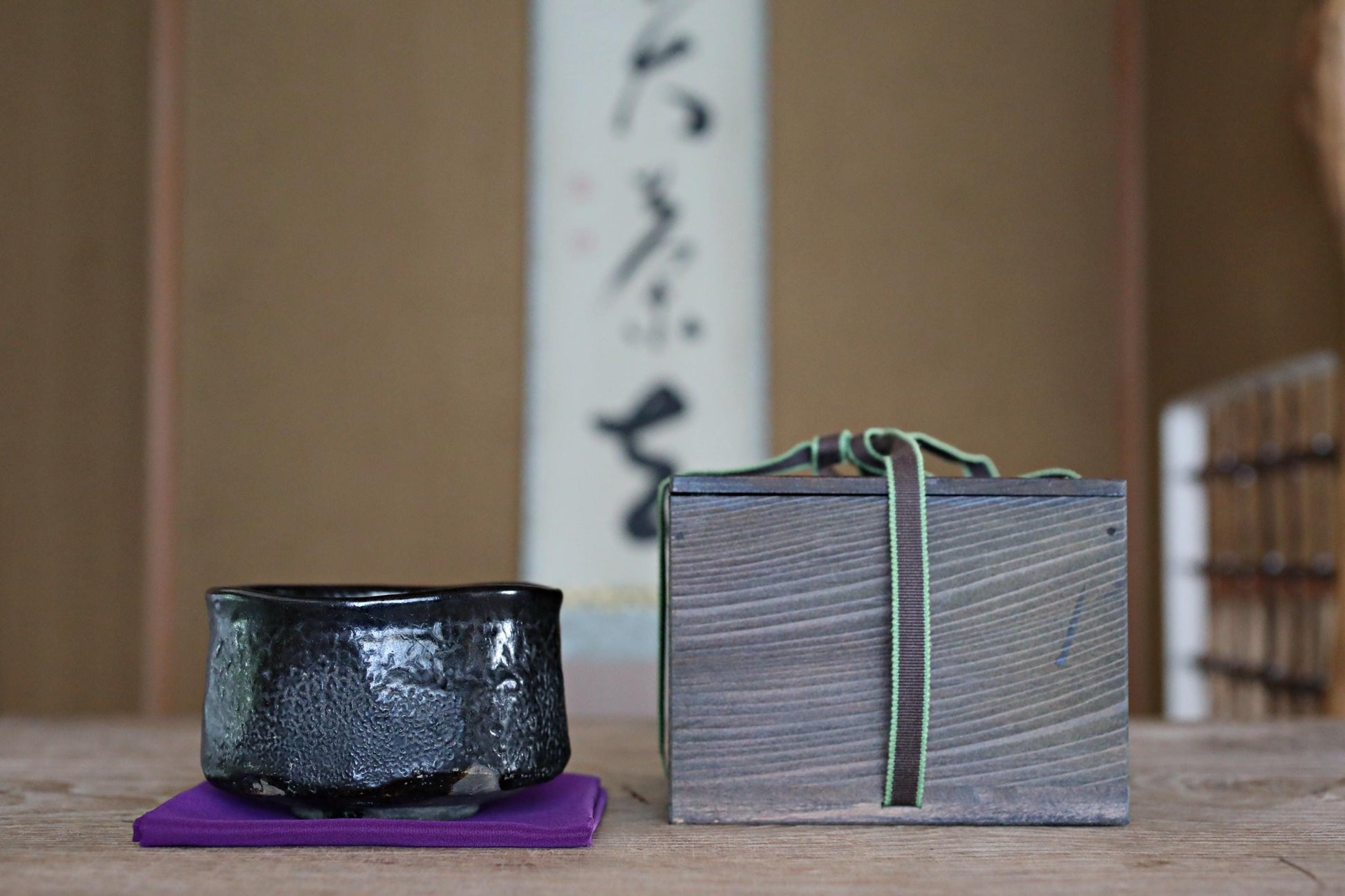 a wooden box and a black ceramic bowl placed on a purple surface, with a blurred background that includes a wall with Japanese characters.
